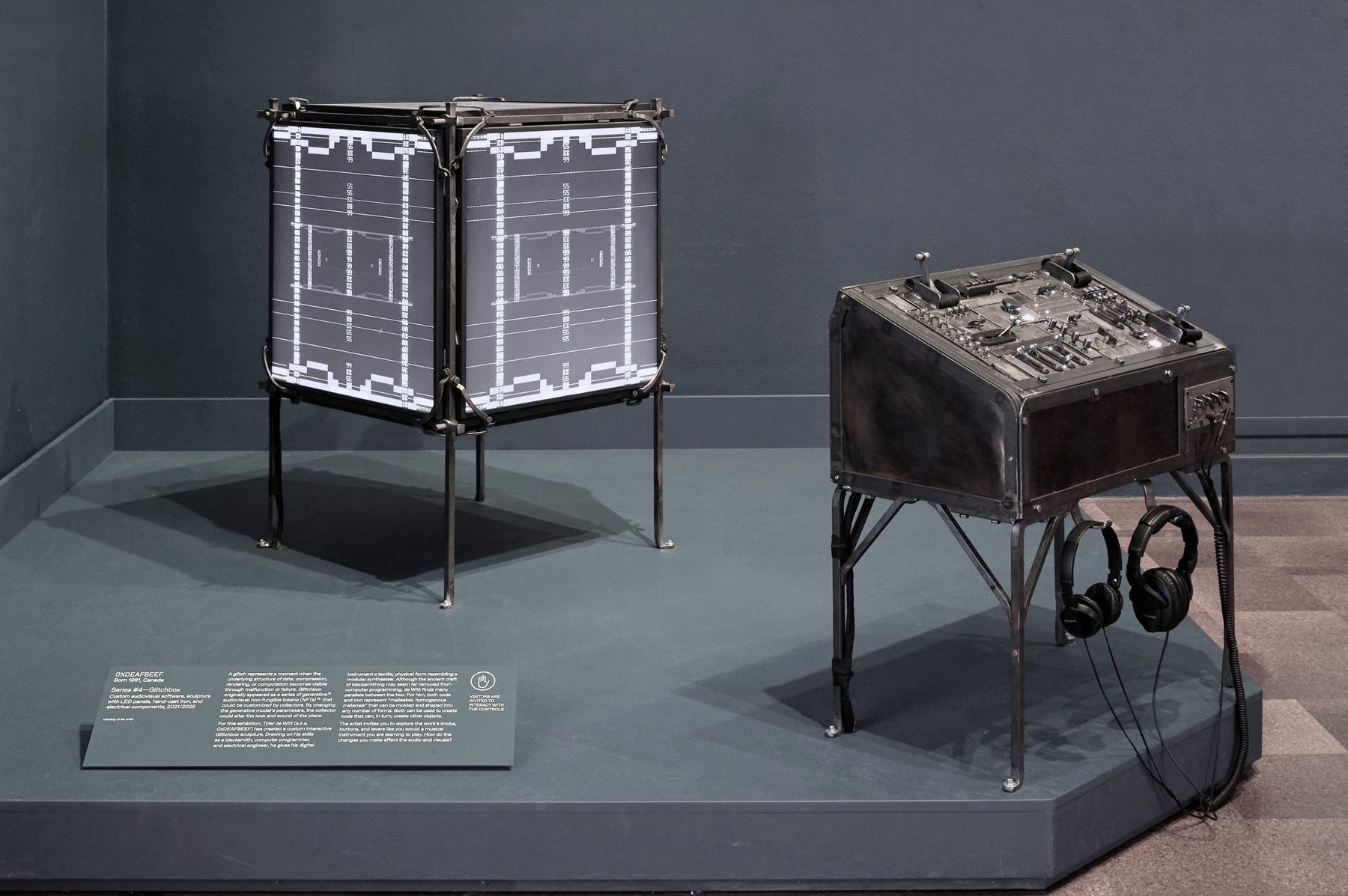 Glitchbox installation — a forged-iron screen cube and a hand-built control console on a plinth in the gallery.